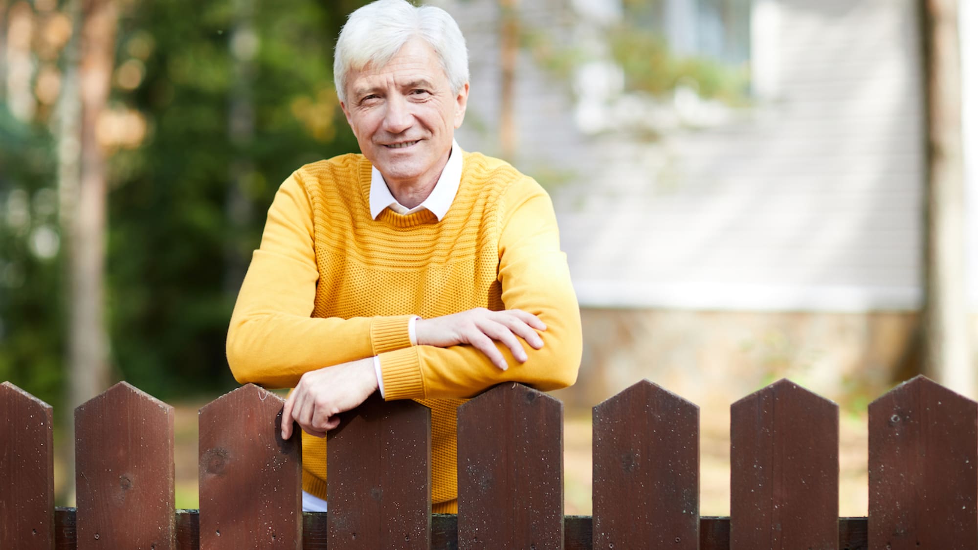 Senior man with grey hair leaning against wooden fence while looking at camera